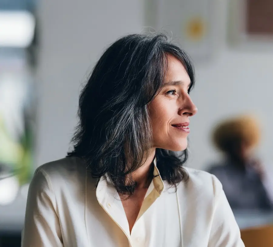 A confident, middle-aged woman wearing professional attire is captured in a bright modern workspace, exuding a sense of sophistication and serenity while looking towards the future with an optimistic expression.