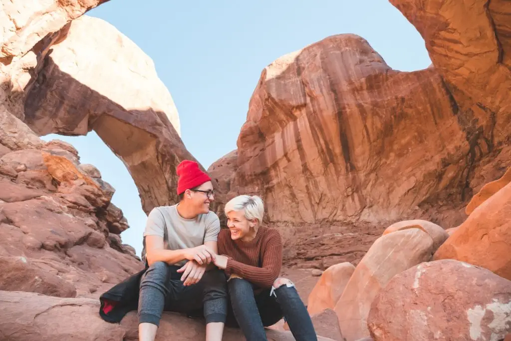 A young couple get up at sunrise to enjoy the majestic view at Arches National Park. They are sitting on a boulder and snuggled up and laughing together. They are surrounded by natural rock formations and a rock bridge in the background.
